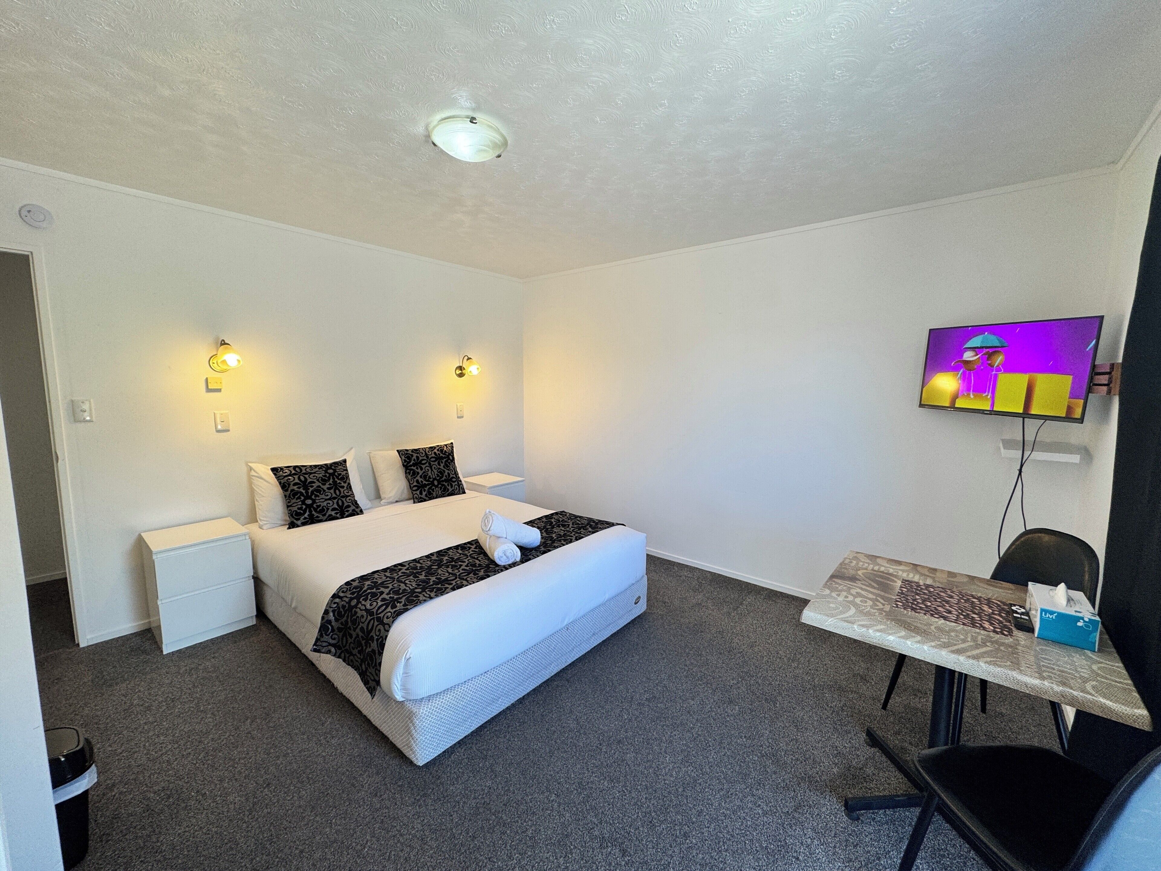 A hotel room with a bed, two bedside tables, a desk with a chair, a television, and a ceiling light.