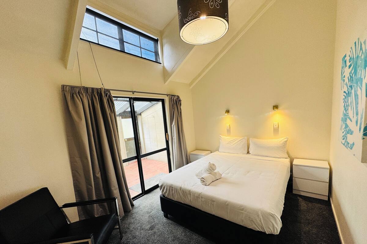 Une chambre d’hôtel comprenant un lit, deux lampes de chevet, une chaise, une fenêtre avec des rideaux et un luminaire au plafond.