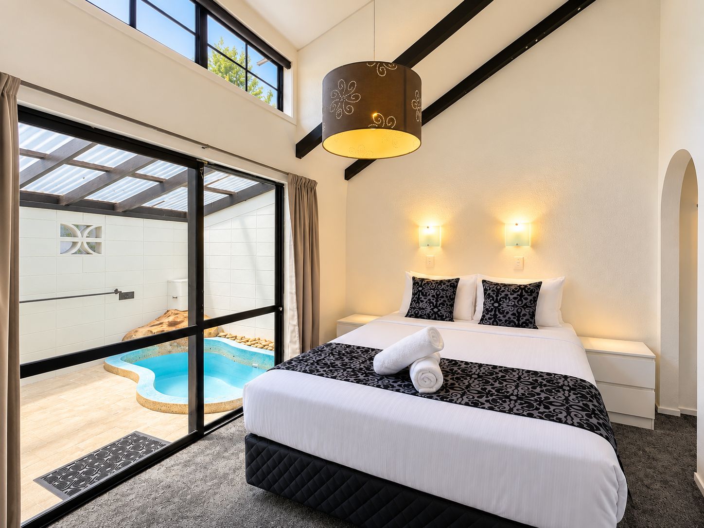 Un dormitorio con una cama grande, dos luces empotradas en la pared, una lámpara de techo, un ventanal corredero que da a una piscina y vistas al exterior.