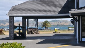 A hotel entrance with a covered patio, a sign encouraging visitors to enjoy New Zealand, and a view of the surrounding landscape.