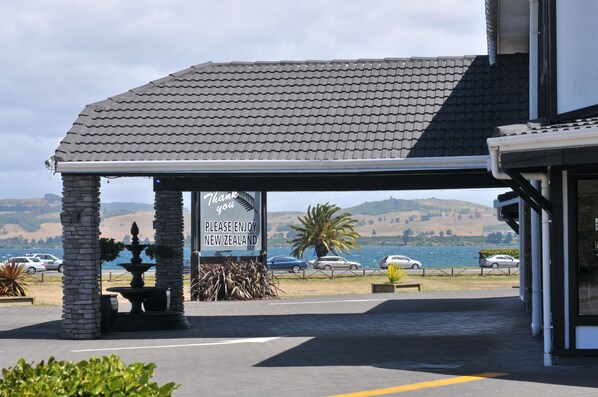 A hotel entrance with a covered patio, a sign encouraging visitors to enjoy New Zealand, and a view of the surrounding landscape.