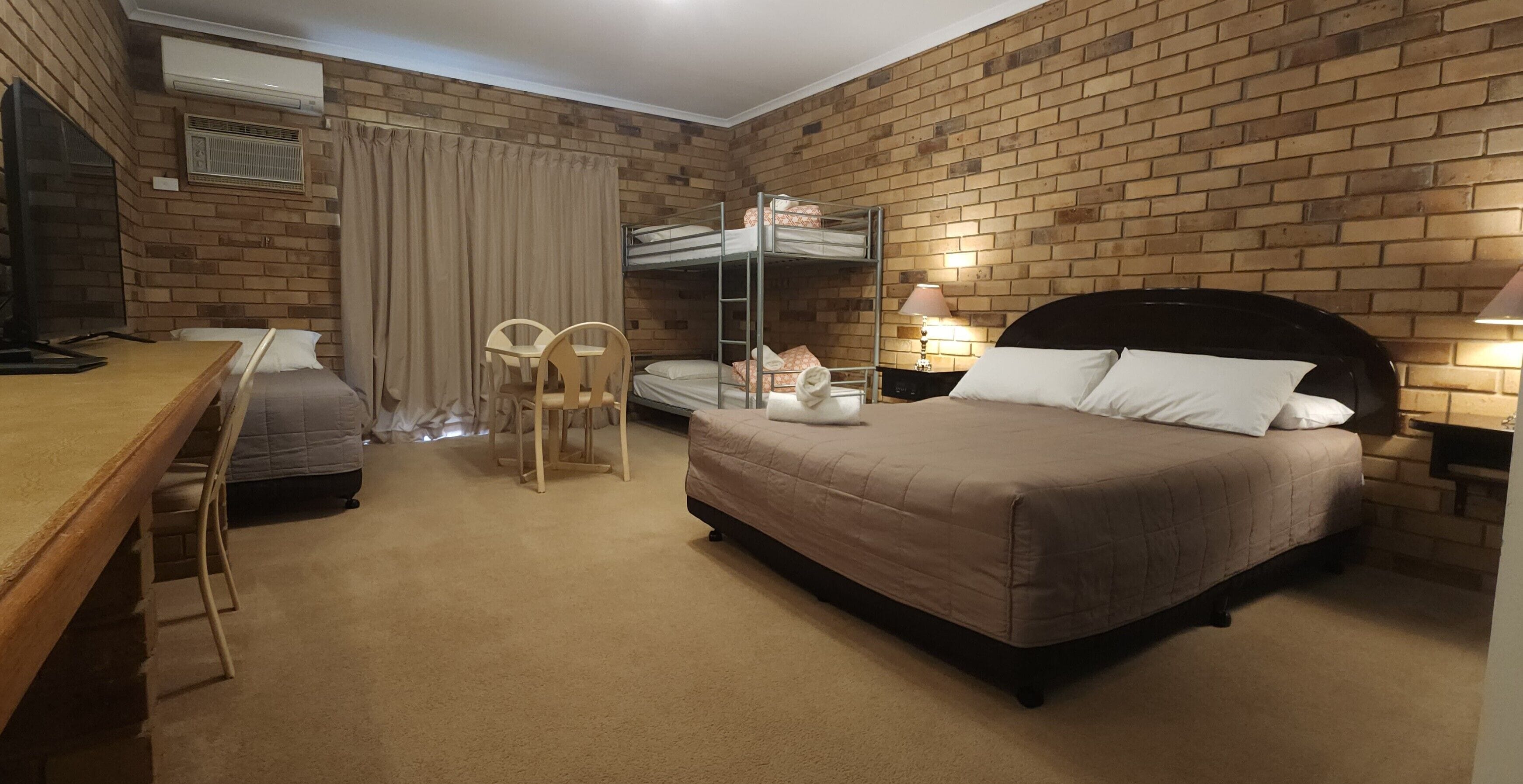 A hotel room with a brick wall, a bunk bed, a desk with a chair, and a bedside table with lamps.