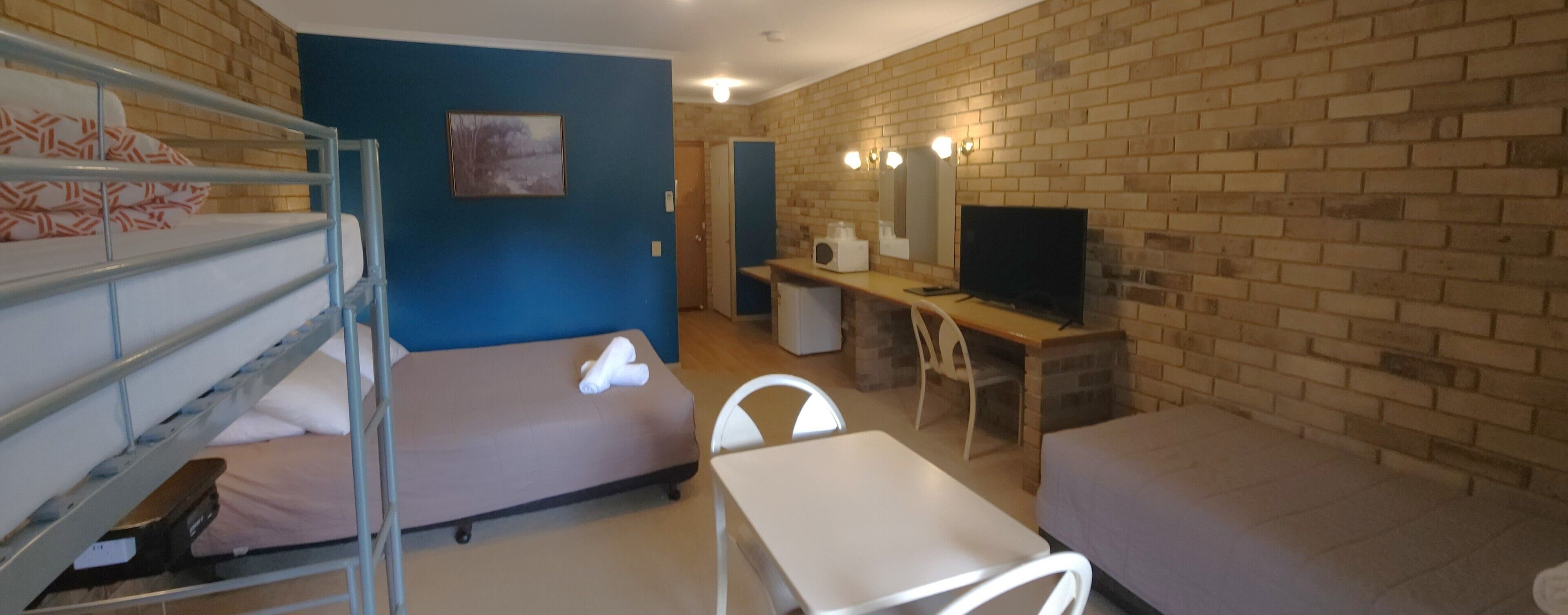 A dormitory room with bunk beds, a desk, a chair, a television, and a small kitchenette.