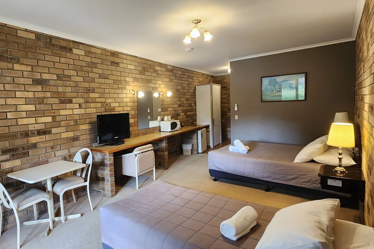 A compact hotel room with a brick wall, a desk with a chair, a bed with pillows, a bedside lamp, a television, and a microwave.