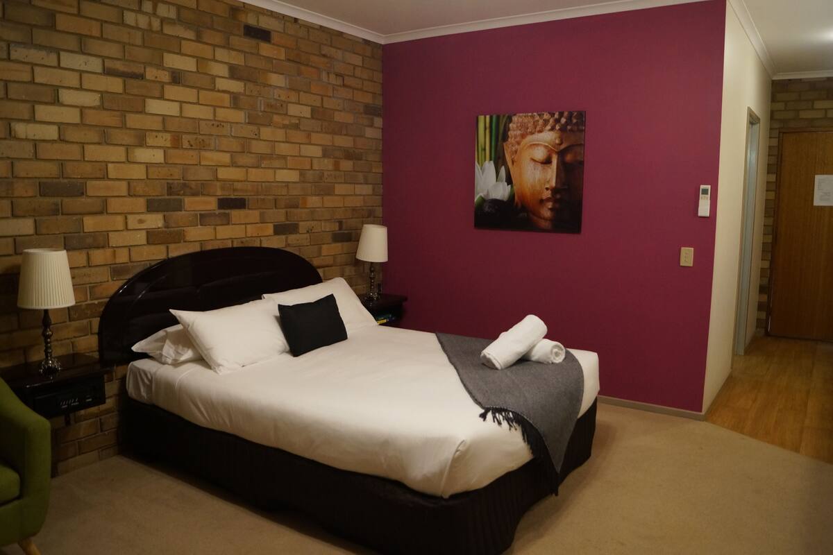 A hotel room with a brick wall, a large bed with white bedding, a purple accent wall, and a Buddha painting.