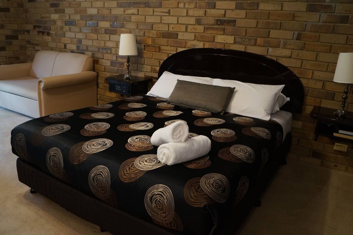 A hotel room with a brick wall, a black bed with a circular pattern, a beige armchair, and two bedside tables with lamps.