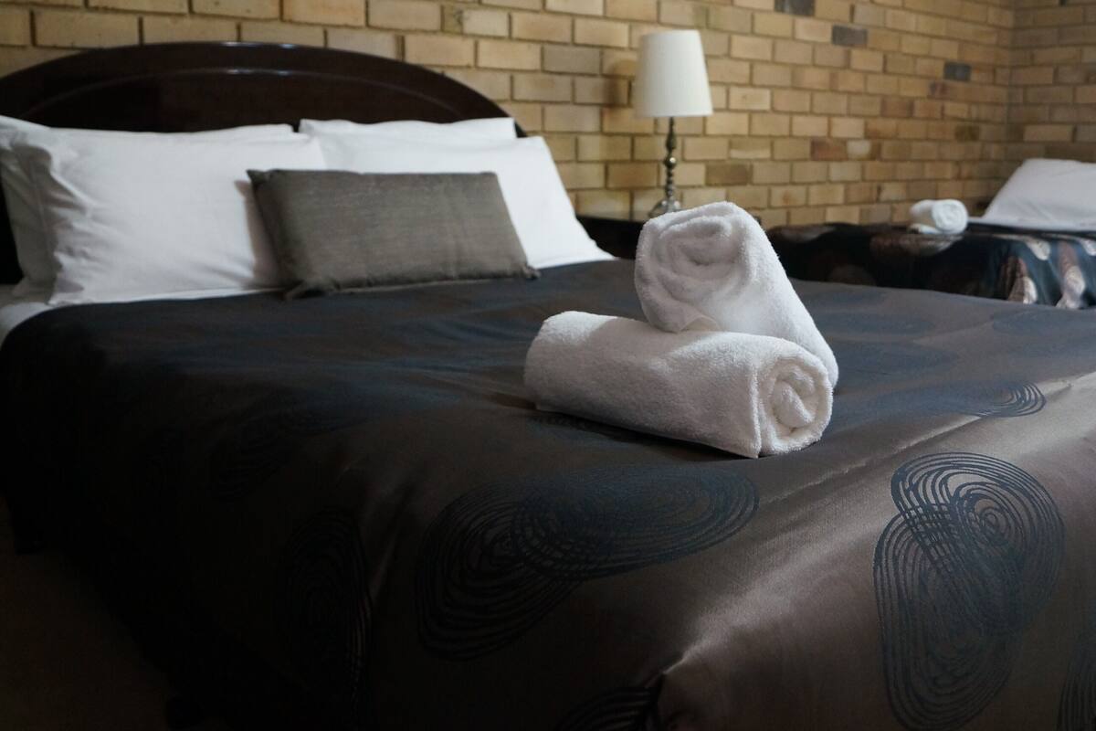 A neatly made bed with a dark patterned bedspread, white pillows, and a folded white towel on the bed.