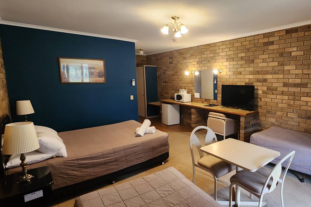 A compact hotel room with a single bed, a desk with a television, a small kitchenette, and a seating area.