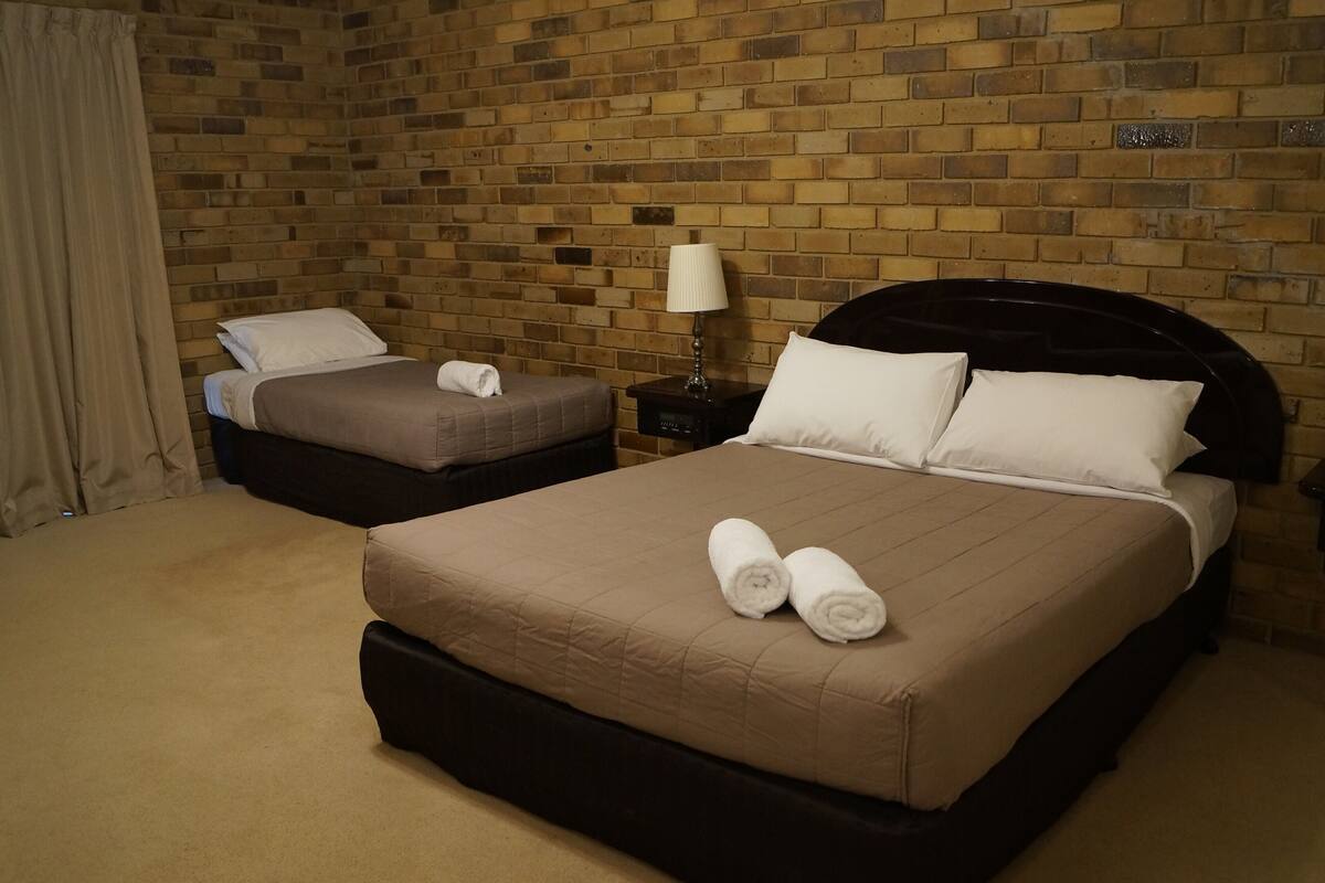 A hotel room with a brick wall, two beds, a nightstand with a lamp, and a curtain.