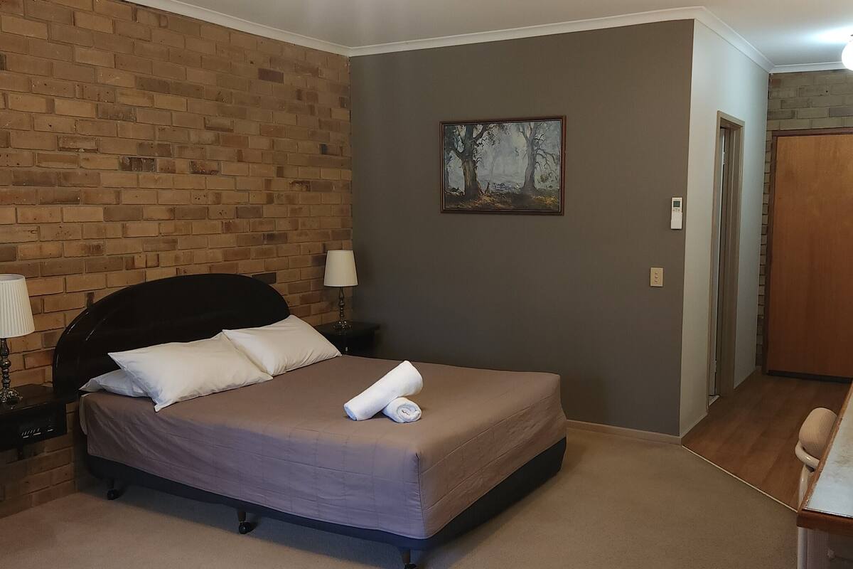A hotel room with a brick wall, a bed with a headboard, a desk with a chair, a painting on the wall, and a door leading to another room.