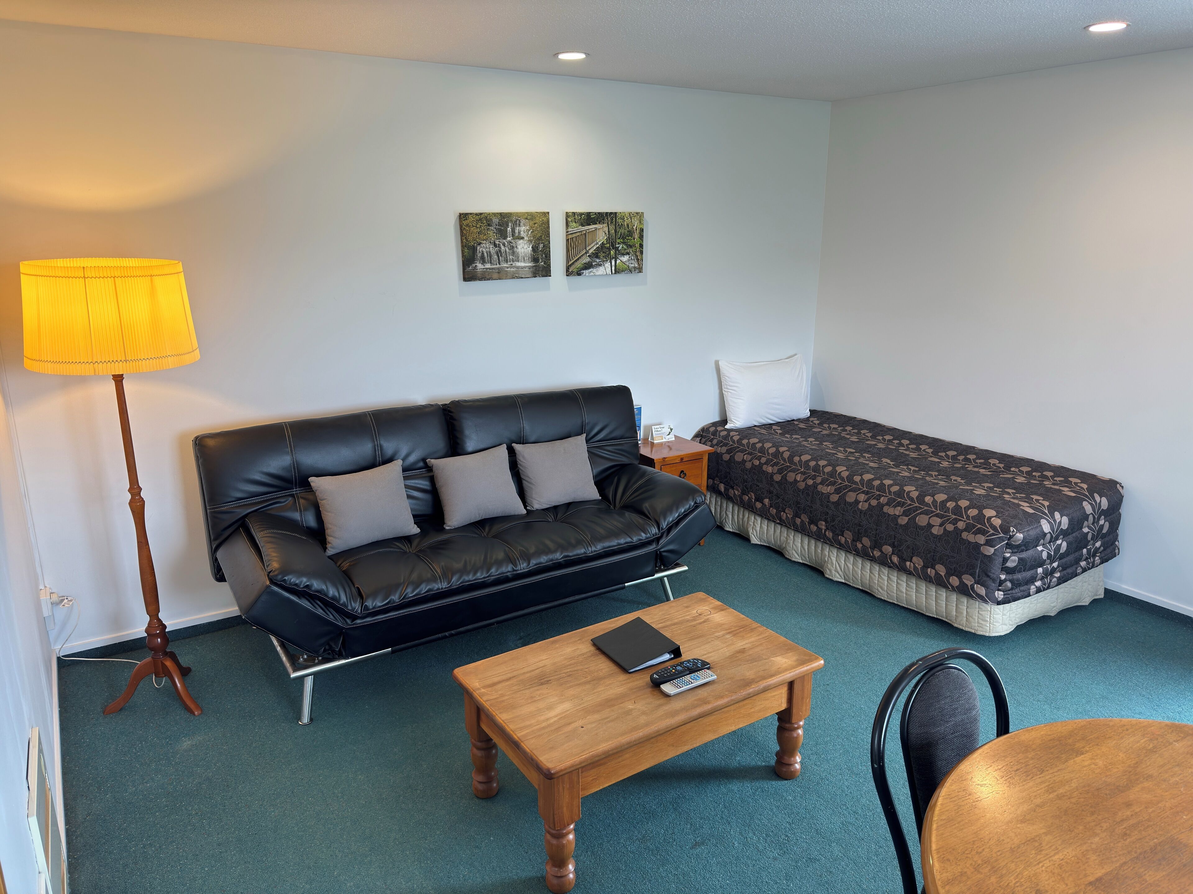 A room with a black leather sofa, a wooden coffee table, a bed with a patterned bedspread, a nightstand, a lamp, and two wall-mounted pictures.