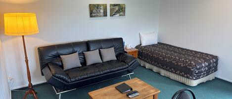 Una habitación con un sofá de cuero negro, una mesa de centro de madera, una cama con colcha estampada, una mesita de noche, una lámpara y dos cuadros colgados en la pared.