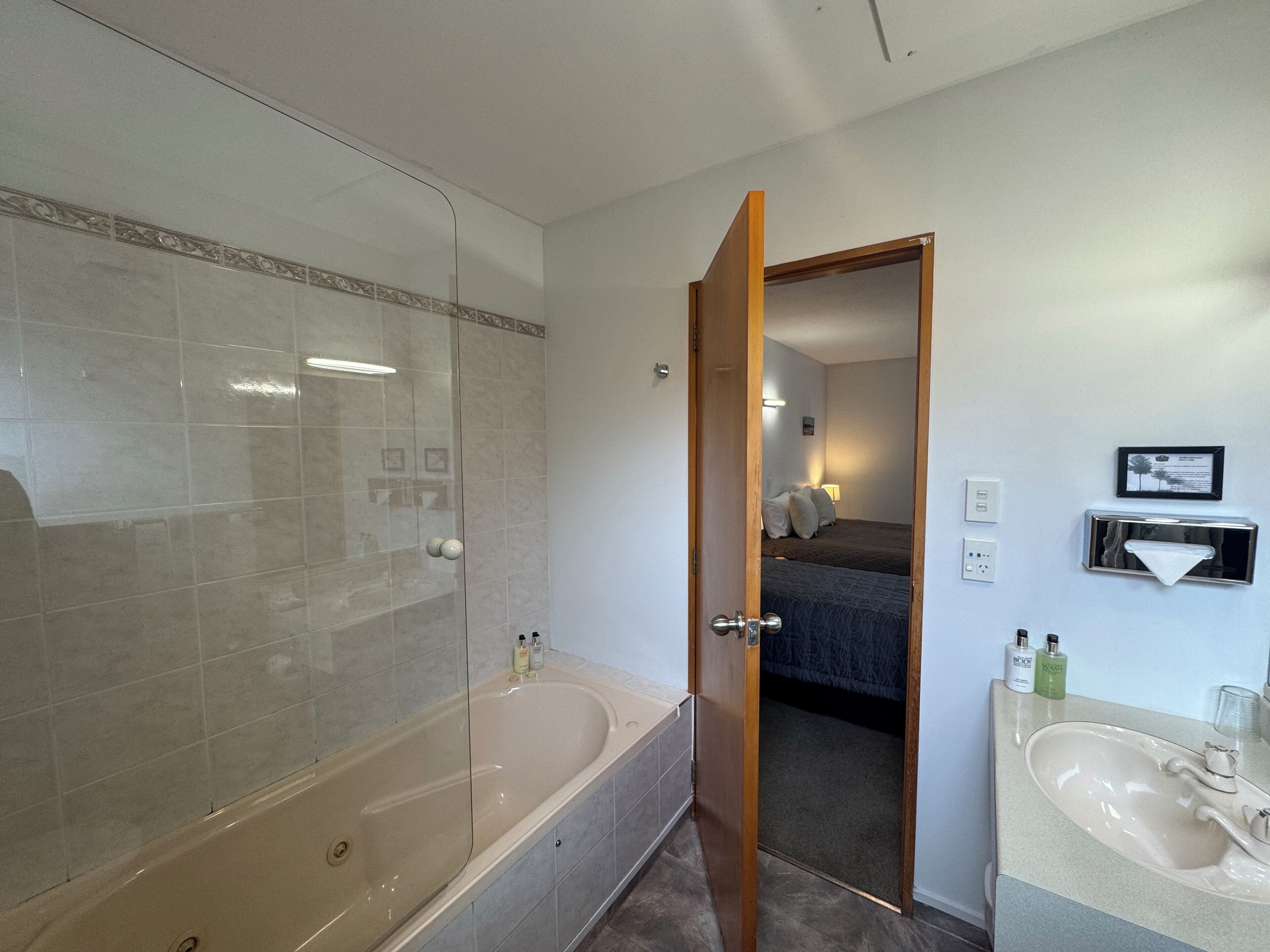 A bathroom with a bathtub, a sink, and a mirror. There is a door leading to a bedroom with a bed and a nightstand.
