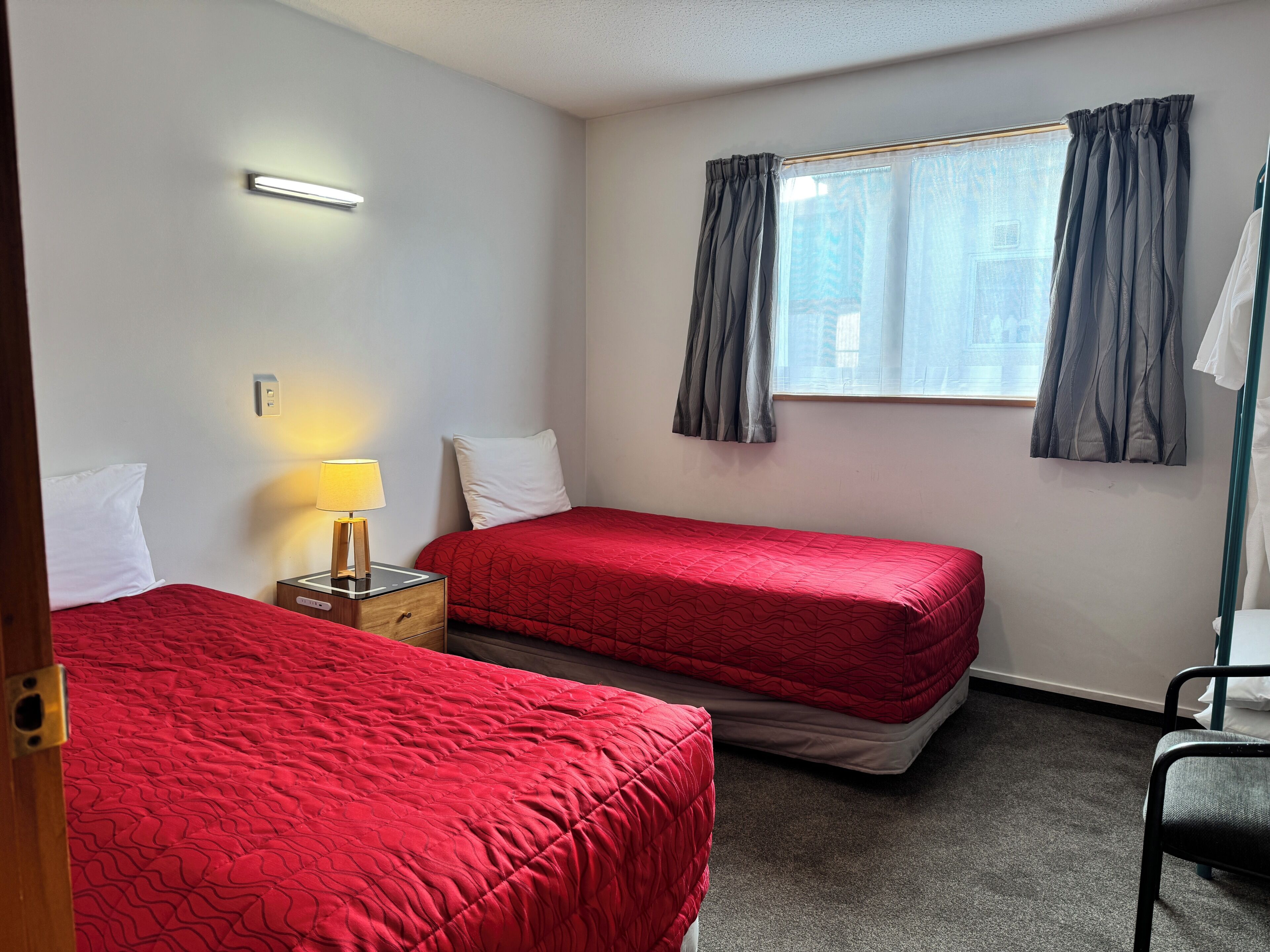 A hotel room with two beds, a nightstand with a lamp, a window with curtains, and a chair.