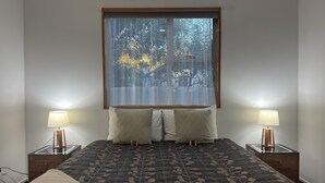 Un dormitorio con una cama grande, dos mesitas de noche con lámparas, un ventanal con cortinas traslúcidas y vistas a árboles.