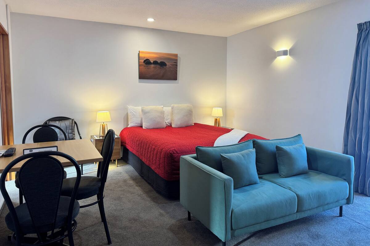 A hotel room with a bed, a desk, a chair, a sofa, and a painting on the wall.