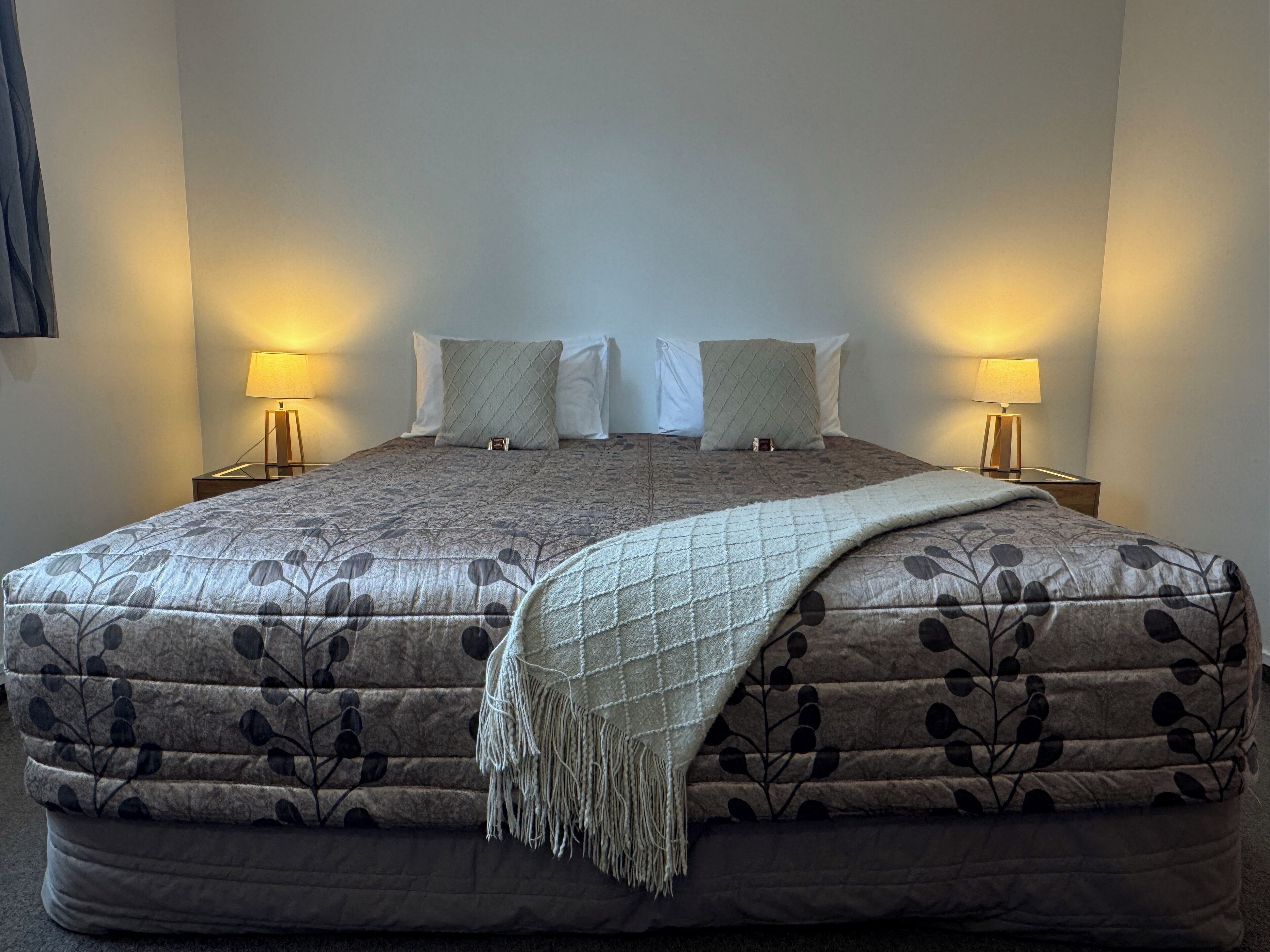 Una cama bien hecha con una colcha estampada y almohadas, con dos lámparas de noche a cada lado sobre mesitas a juego.