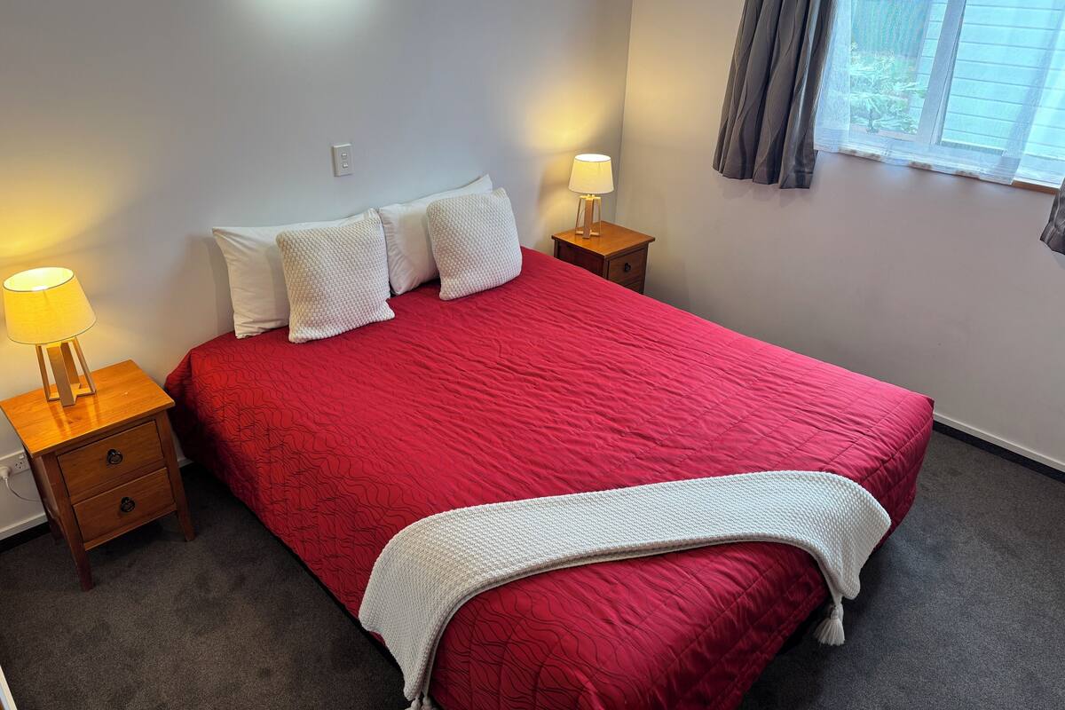 A neatly made bed with a red bedspread, two white pillows, a white throw blanket, and two bedside lamps on wooden nightstands.