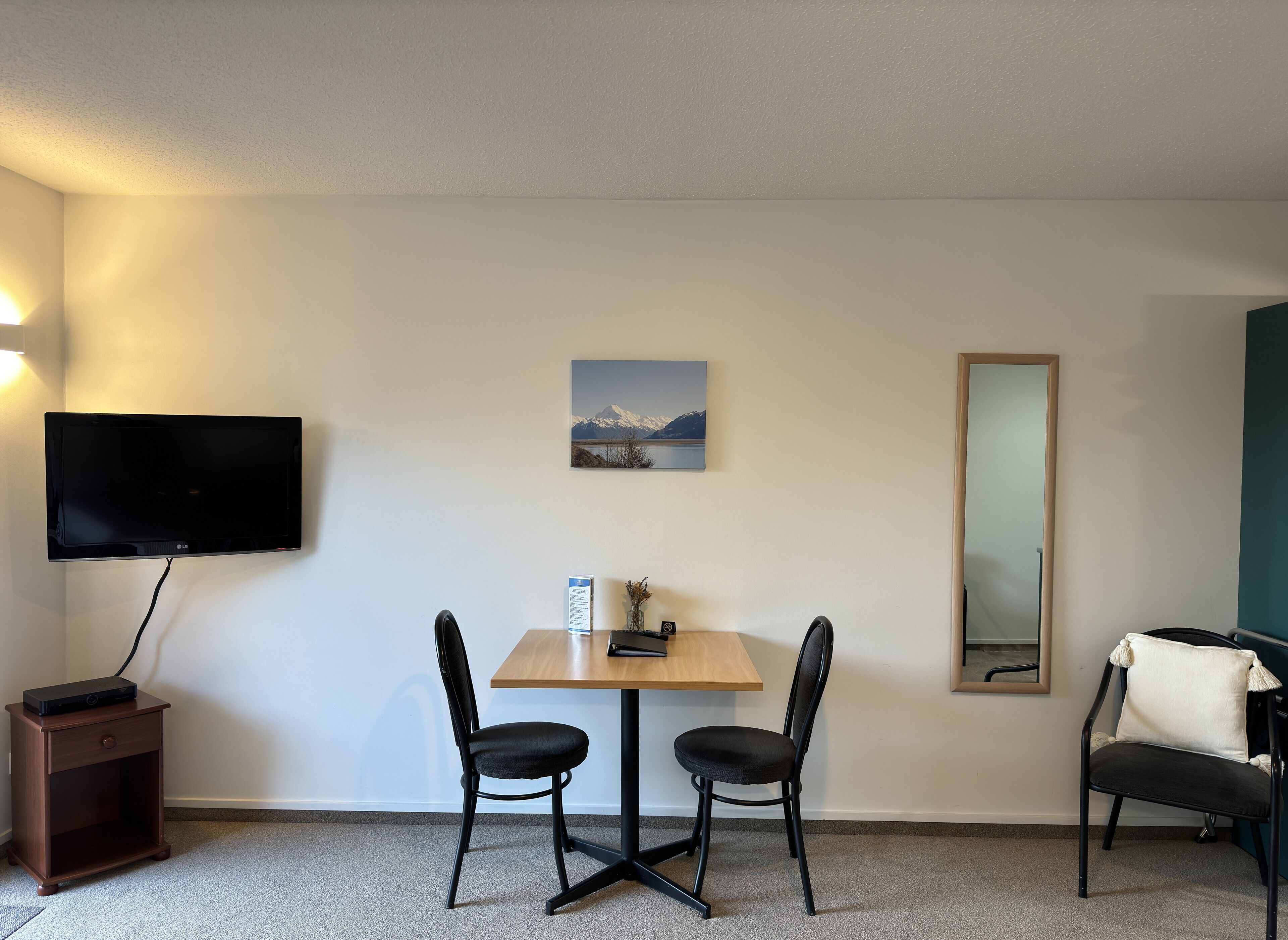 A hotel room with a flat-screen TV mounted on the wall, a small table with two chairs, a wooden nightstand, a mirror, and a framed picture of a mountain landscape.