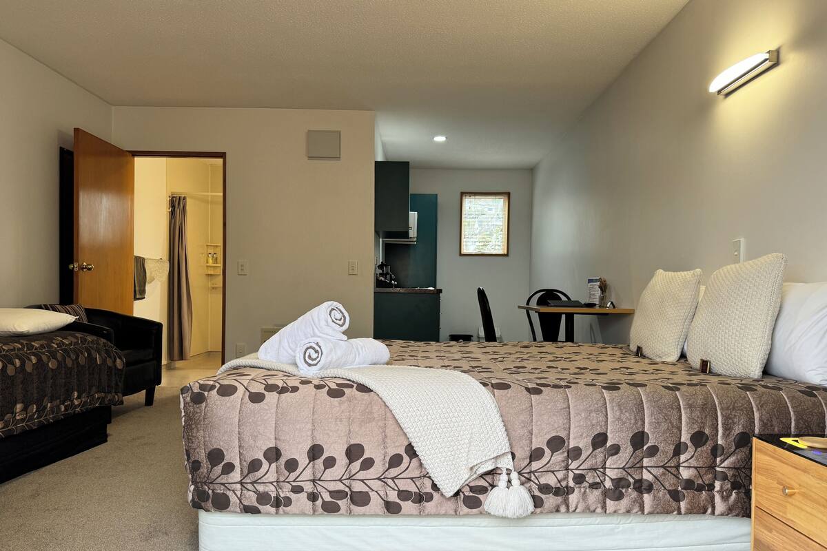 A bedroom with a large bed, a desk, and a chair. There is a door leading to another room.