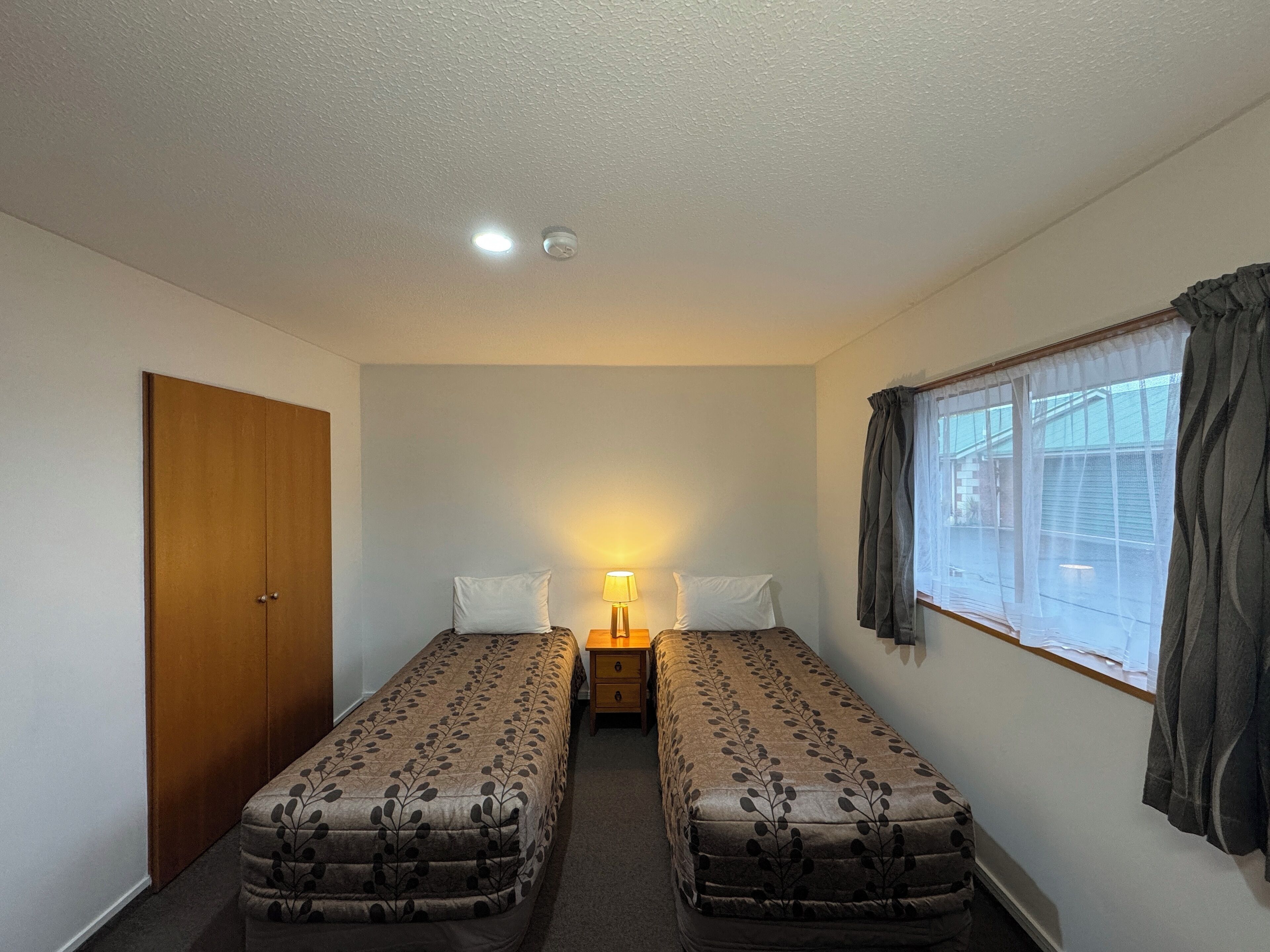 A small, clean room with two single beds, a wooden door, a window with curtains, and a nightstand with a lamp.