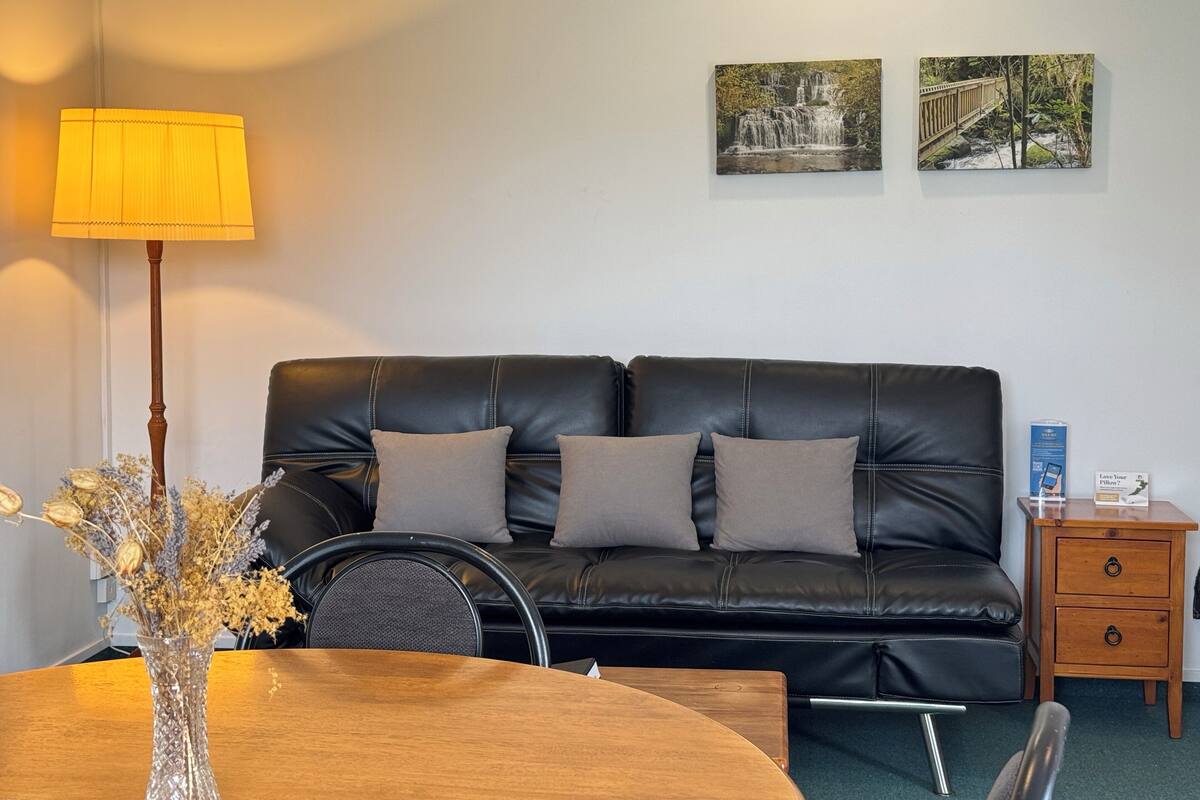 A room with a black leather sofa, a wooden side table, a lamp, a vase with dried flowers, and two framed pictures on the wall.