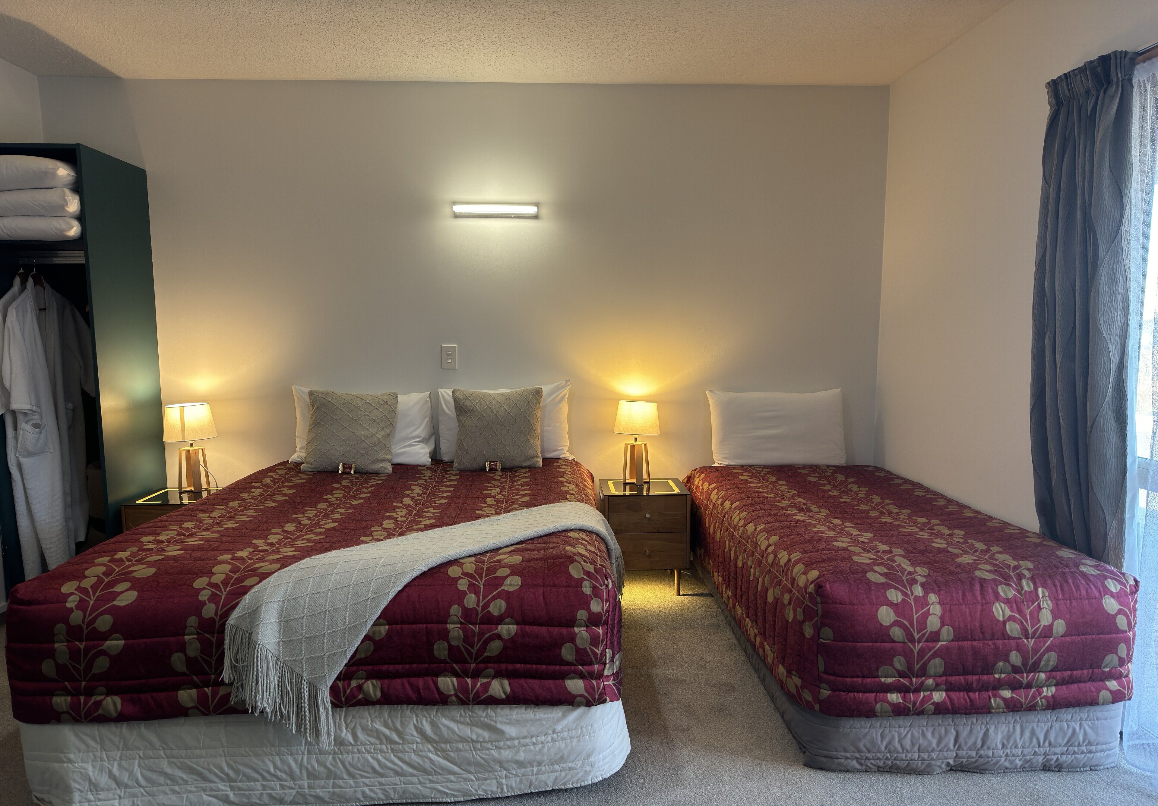 Ett hotellrum med två sängar, var och en med ett rött, blommigt sängöverkast och vita kuddar. Det finns ett nattduksbord med en lampa på varje sida av sängarna.