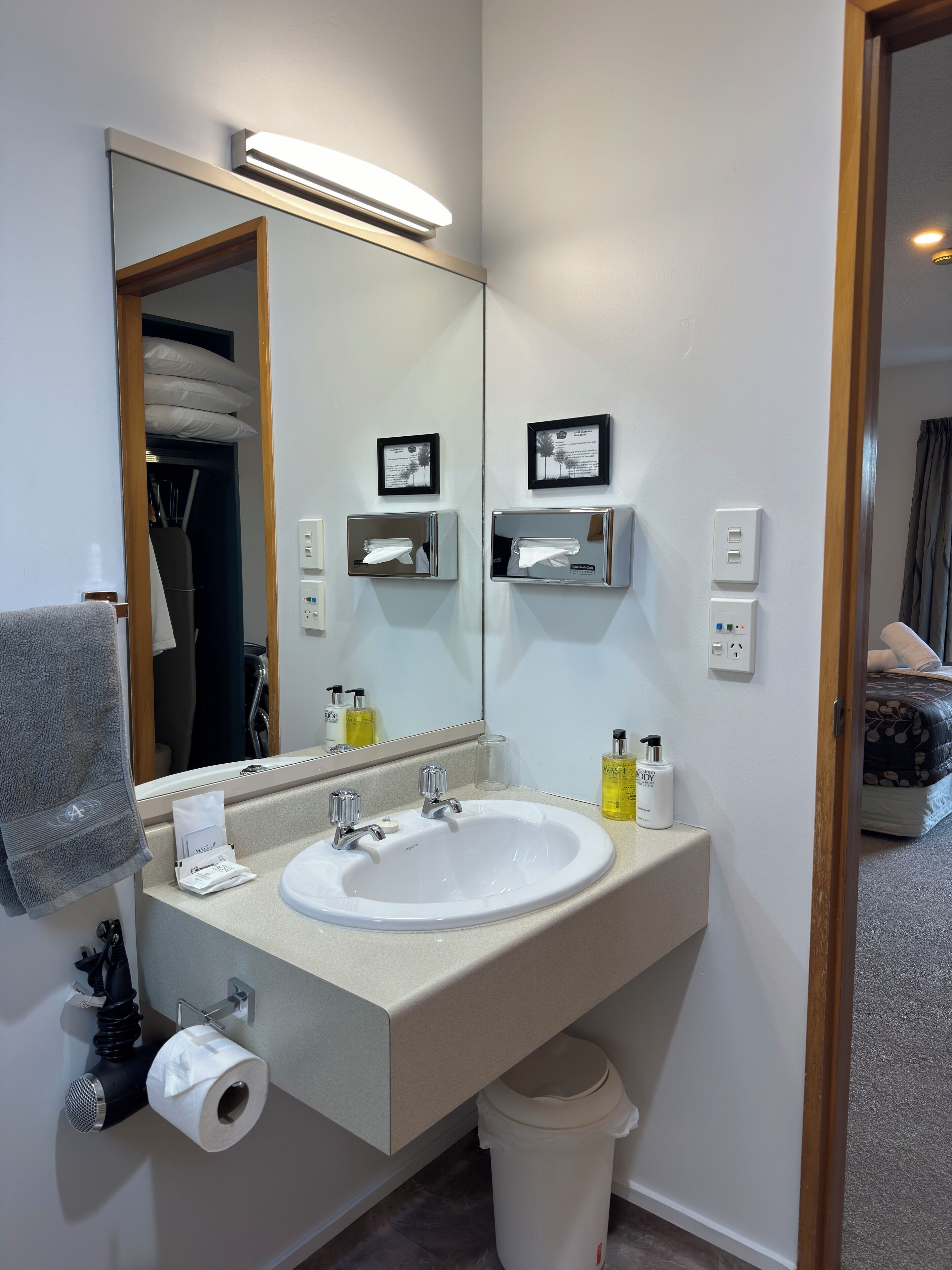 A hotel bathroom with a large sink, mirror, towel rack, and a trash bin.