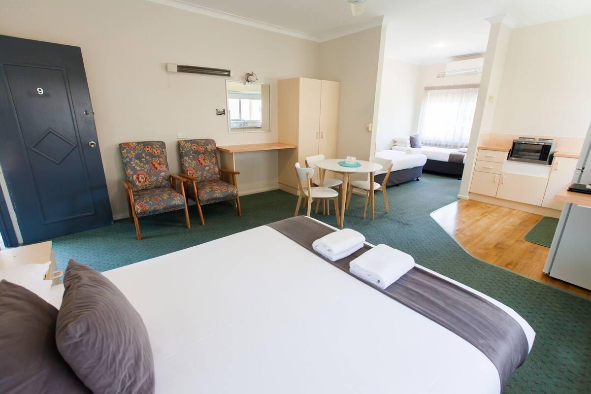 A hotel room with a bed, two armchairs, a desk, a small table, a kitchenette, and a bathroom.