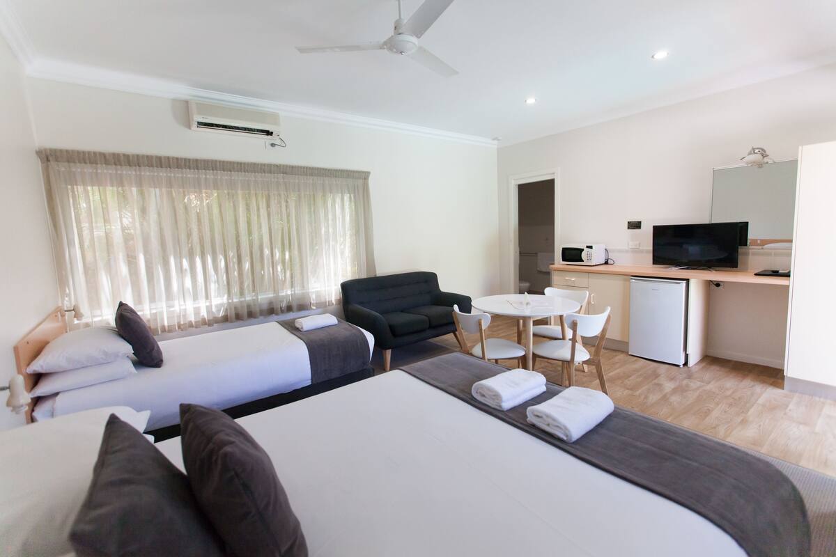 A hotel room with two beds, a desk with a TV, a small fridge, and a dining area.