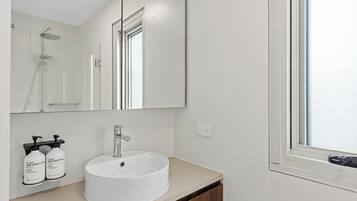 Un baño con un lavamanos blanco, un espejo grande y una ducha. Hay dos botellas de crema para manos y cuerpo sobre el mostrador.