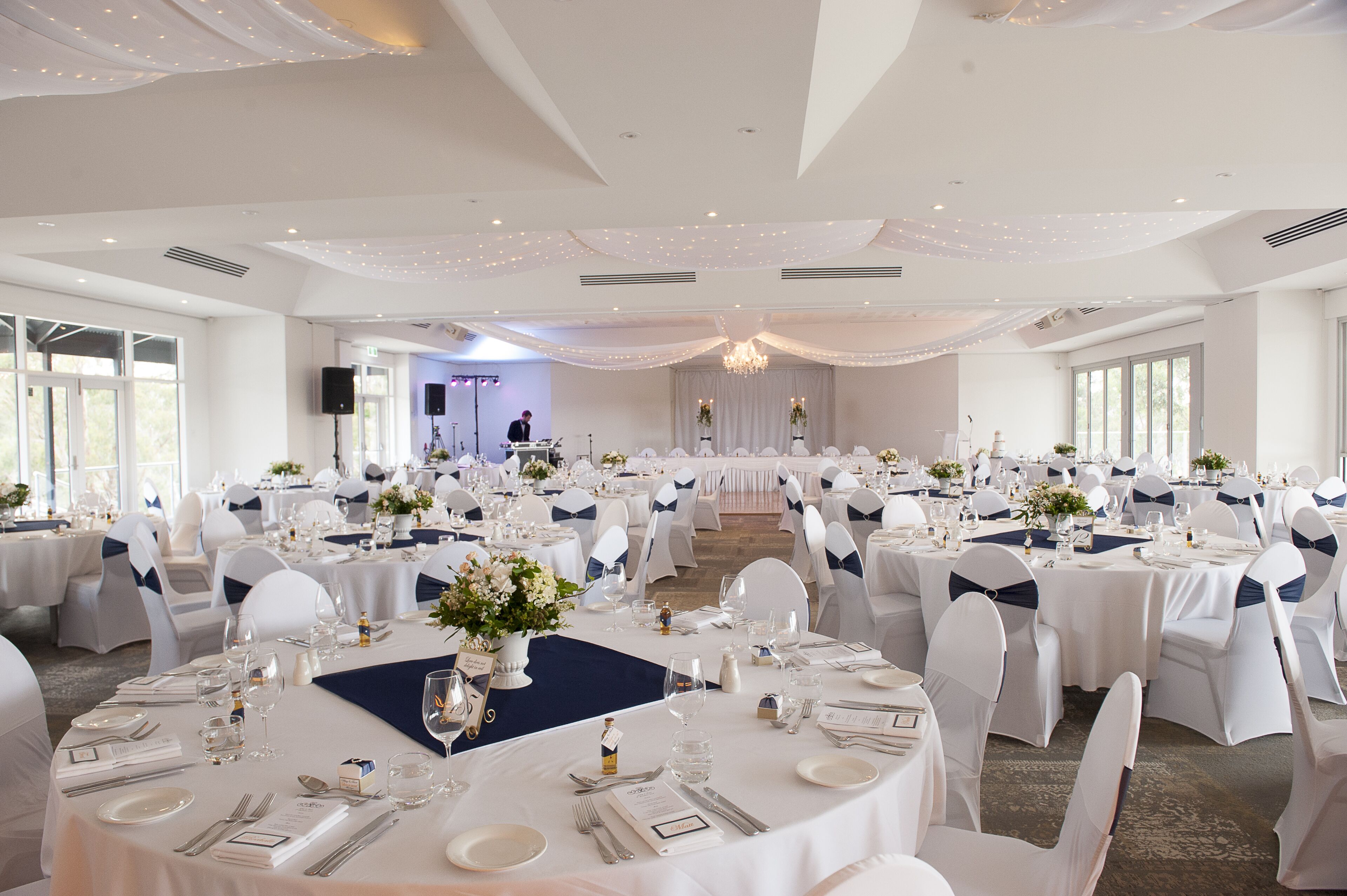 Un salón de banquetes con mesas redondas dispuestas para un evento formal, con manteles blancos, listones azules y centros de mesa florales.