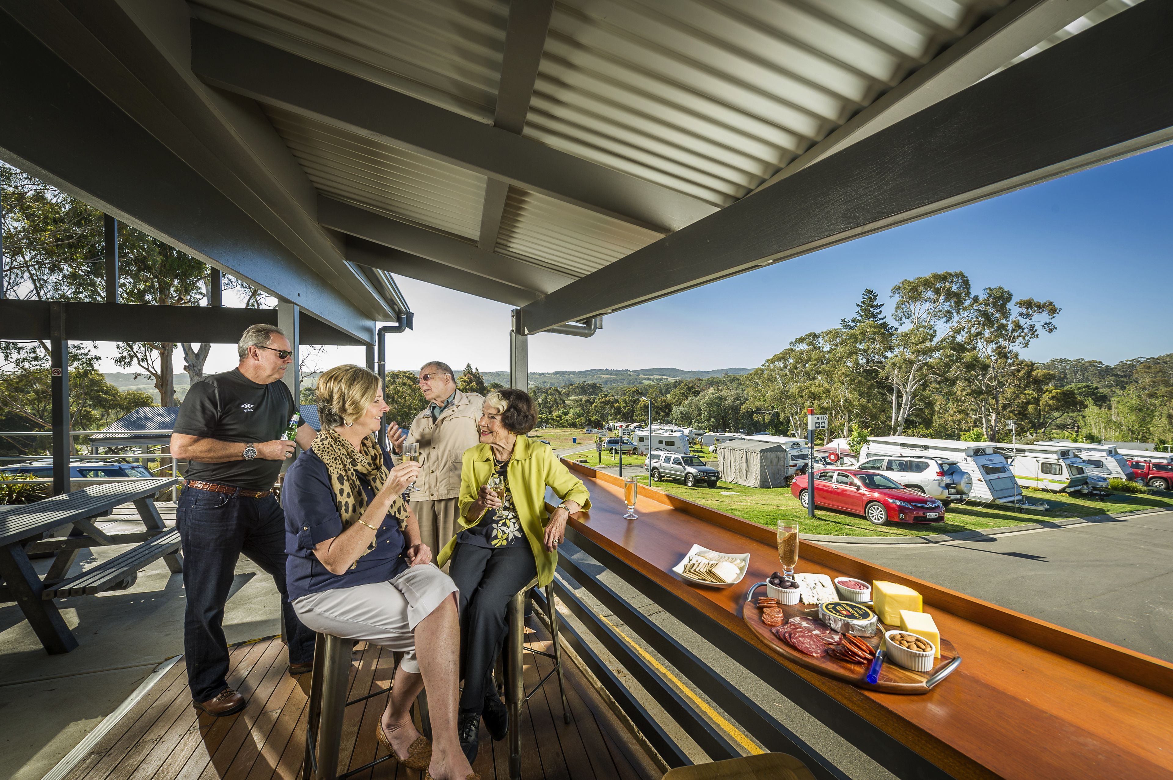 A covered outdoor seating area with a wooden deck, a bar with food and drinks, and a view of a parking lot with RVs.