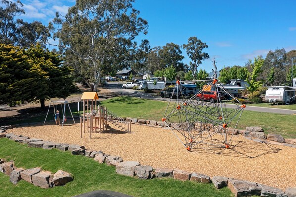 Un parque infantil con una estructura para escalar y columpios, rodeado de una zona de grava y un campo de césped.