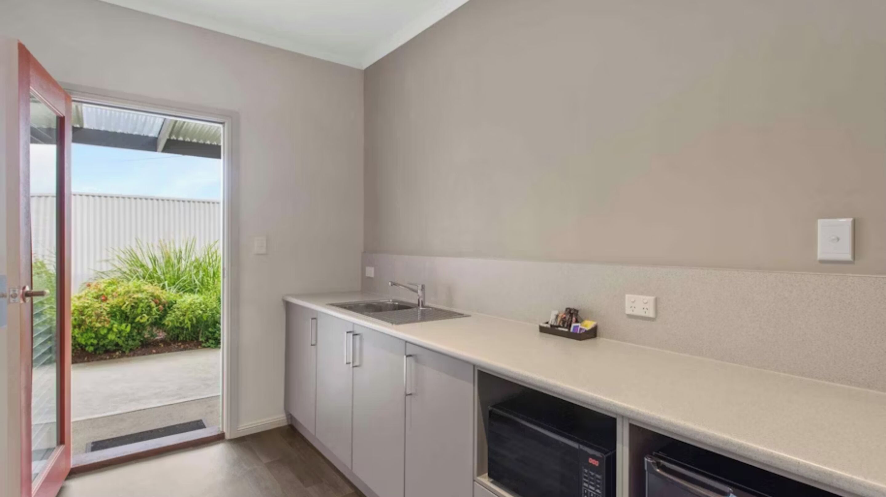 A modern kitchen with a white countertop, a sink, and cabinets. There is a sliding glass door leading to a balcony with greenery.
