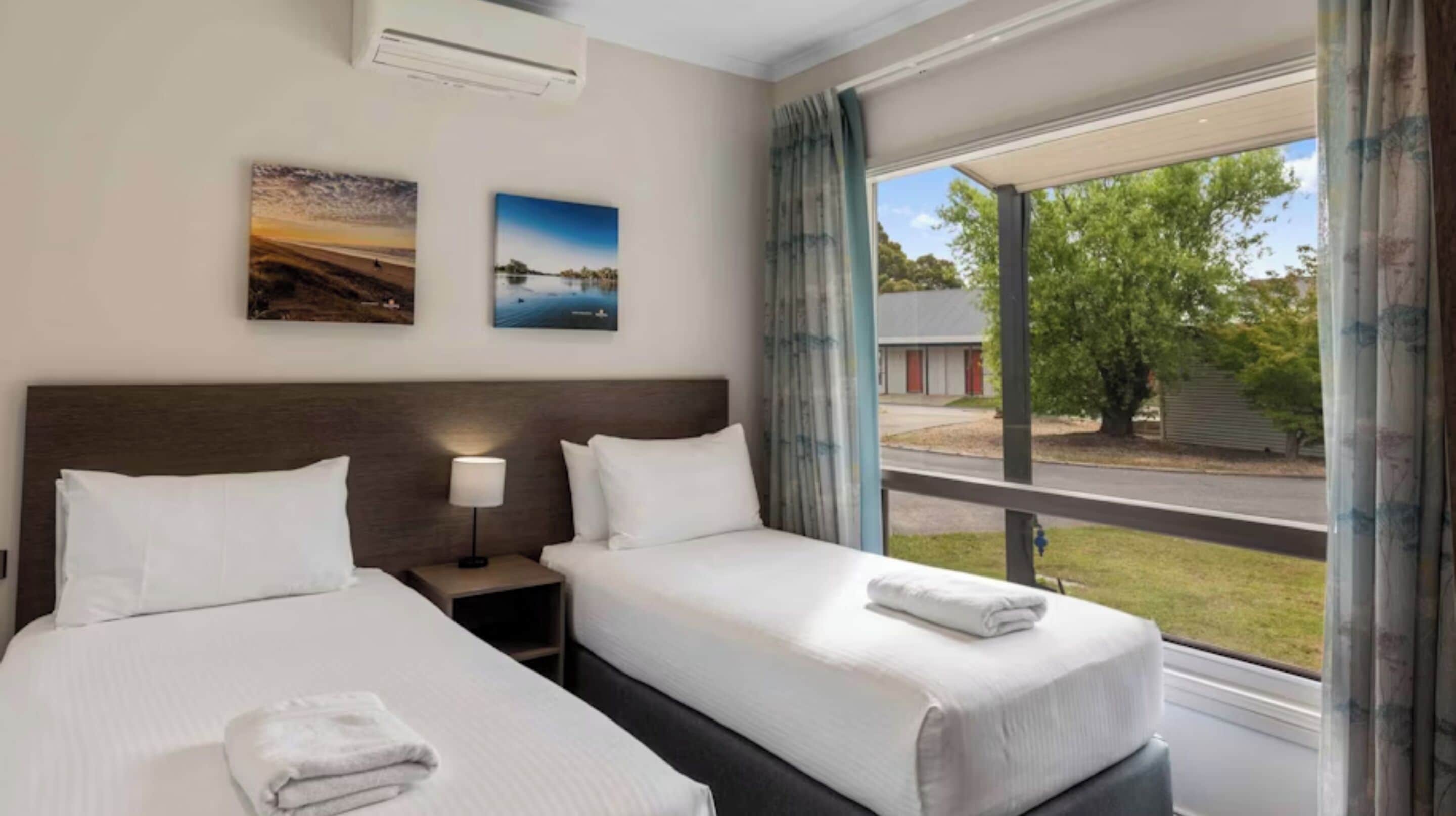 Een hotelkamer met twee bedden, een nachtkastje met een lamp, en een groot raam met uitzicht.