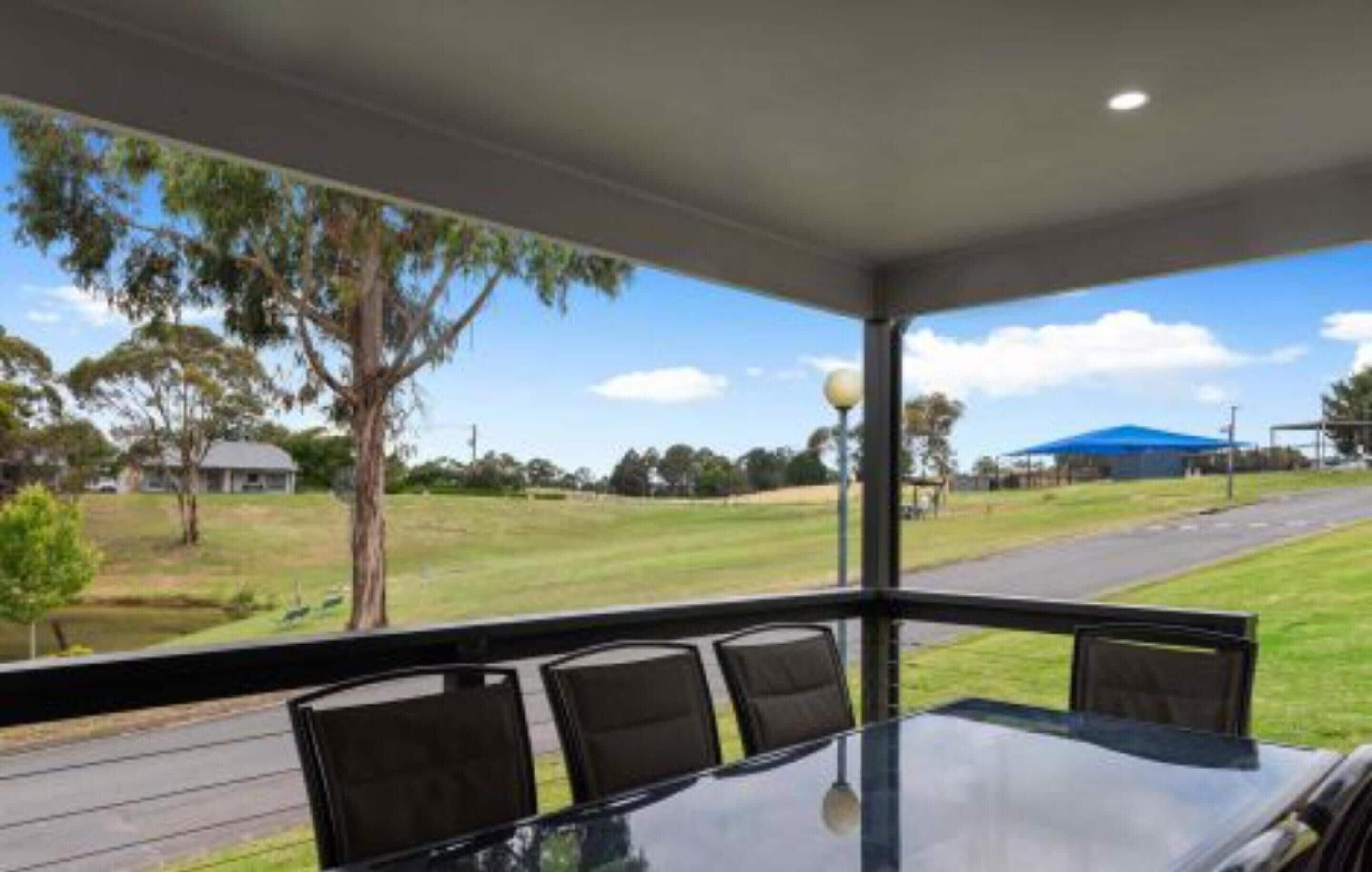A room with a large glass table and four chairs, a view of a grassy area and a road outside.