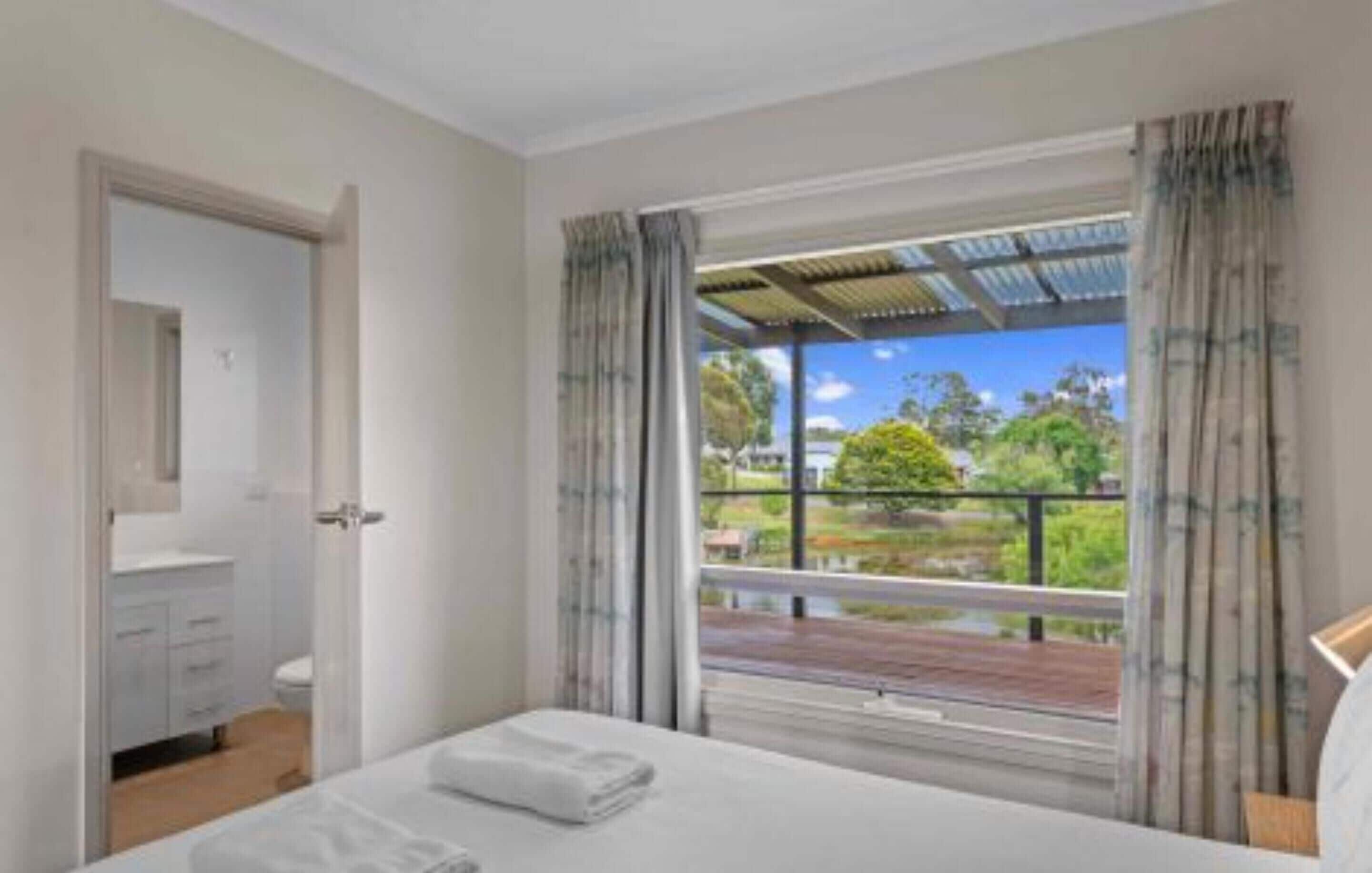 A bedroom with a bed, a bathroom with a toilet and sink, and a balcony with a view of greenery and a building.
