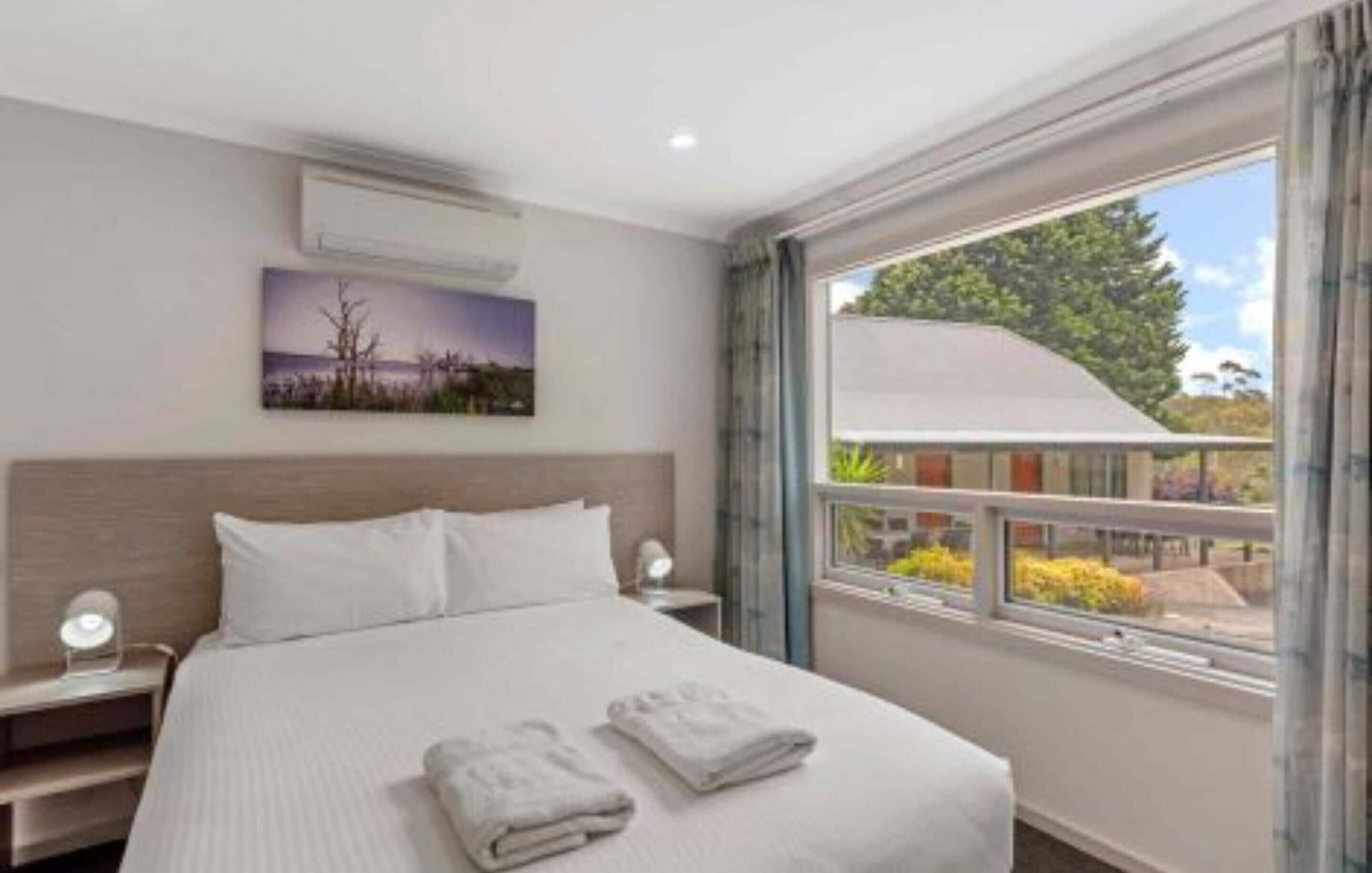 Hotelkamer met een bed, nachtkastje, wandairconditioner, raam met uitzicht op groen en een schilderij aan de muur.