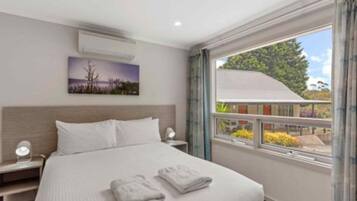 Hotelkamer met een bed, nachtkastje, wandairconditioner, raam met uitzicht op groen en een schilderij aan de muur.