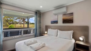 Habitación de hotel con una cama grande, ventana con vistas a la vegetación, equipo de aire acondicionado y mesita de noche con lámpara.