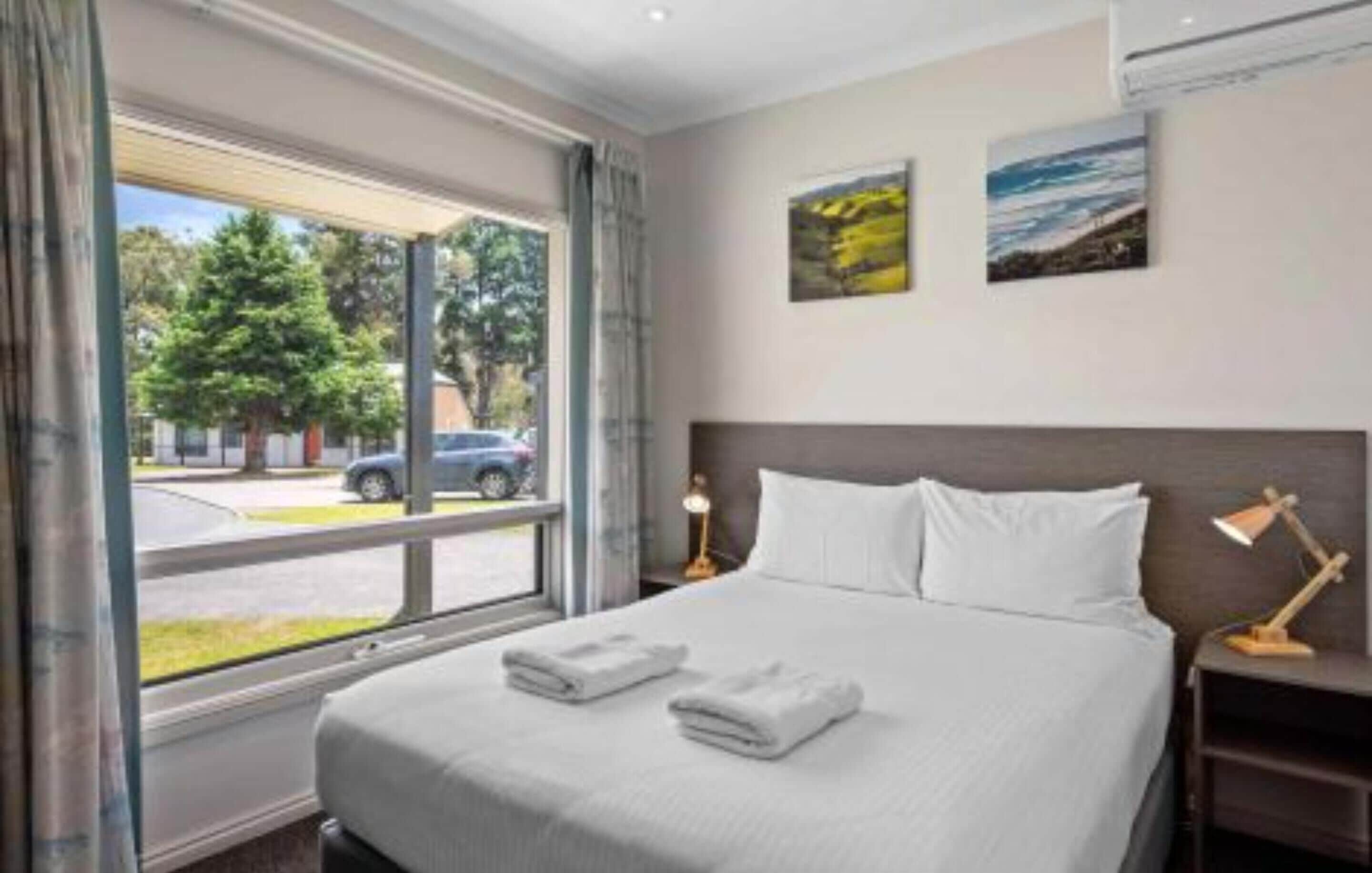 Habitación de hotel con una cama grande, dos almohadas, una mesita de noche con lámpara y una ventana que ofrece vistas a la calle y a los árboles.