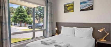 Habitación de hotel con una cama grande, dos almohadas, una mesita de noche con lámpara y una ventana que ofrece vistas a la calle y a los árboles.