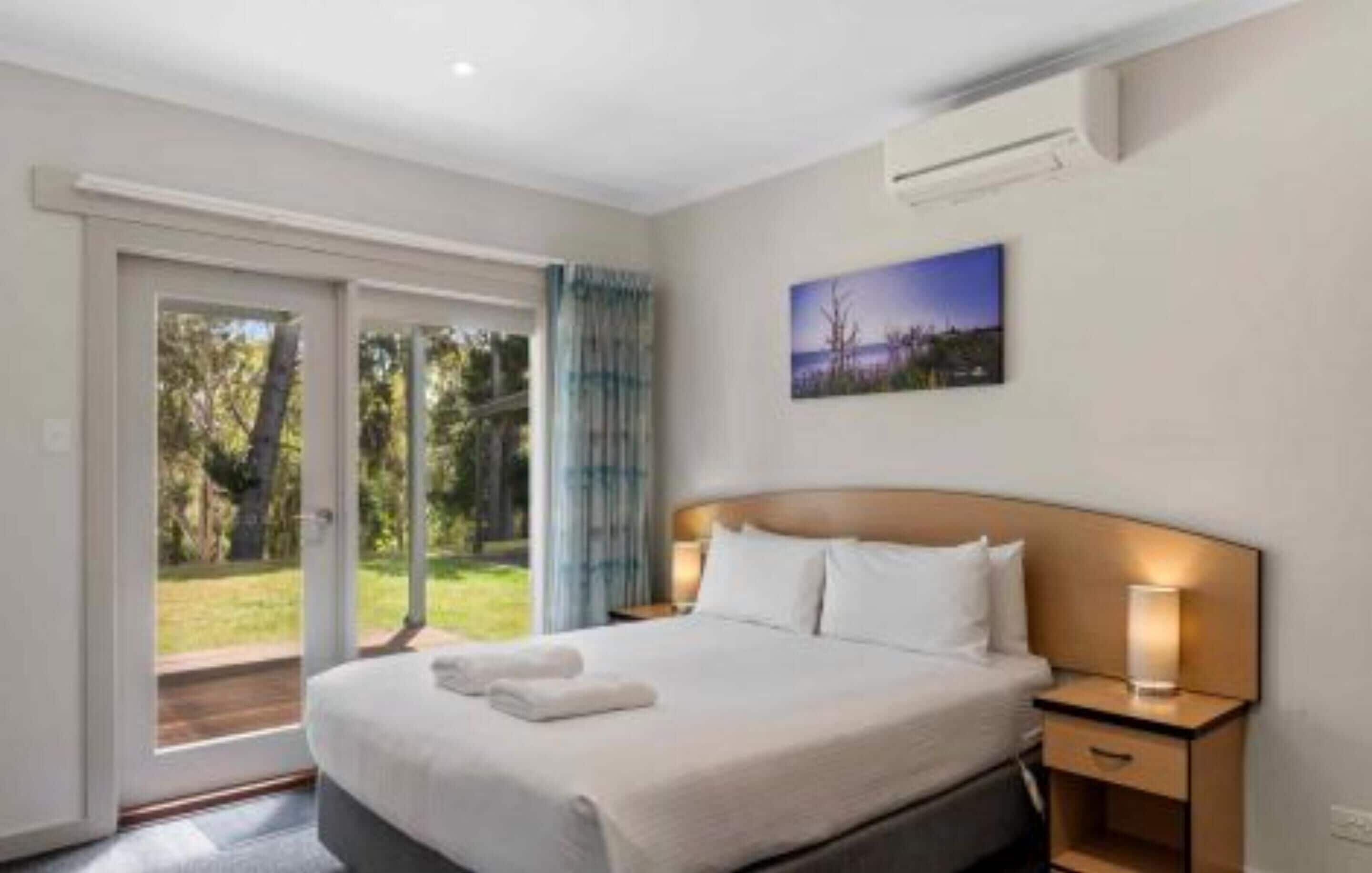 A hotel room with a large bed, a nightstand, and a view through a sliding glass door to an outdoor area.