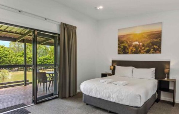 A bedroom with a large bed, a nightstand, a painting of a landscape, and a sliding glass door leading to an outdoor area. - Discovery Parks - Hahndorf (Hahndorf)