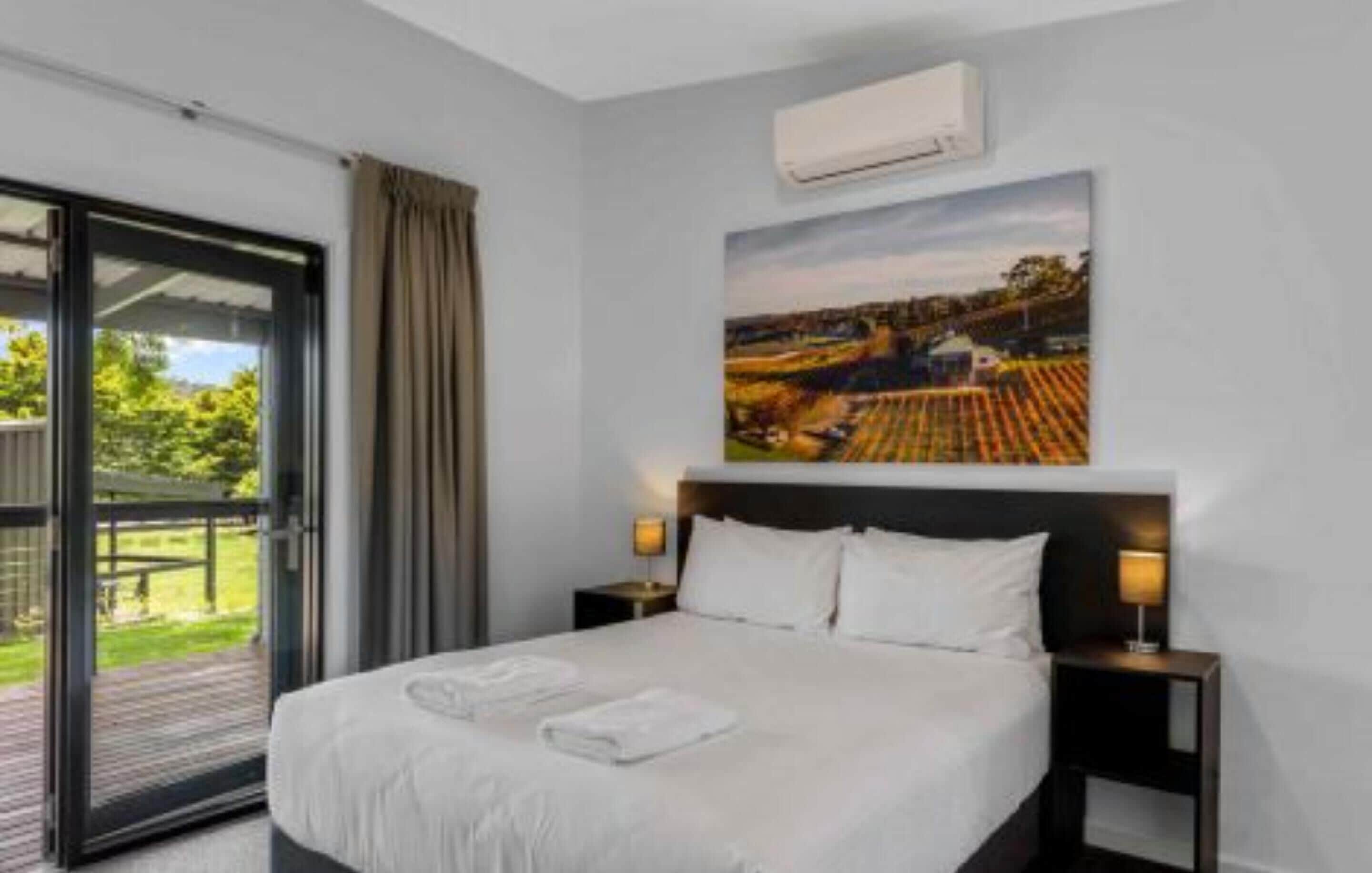 A hotel room with a bed, two bedside tables with lamps, a large window with curtains, and a painting on the wall.