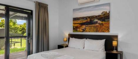 A hotel room with a bed, two bedside tables with lamps, a large window with curtains, and a painting on the wall.