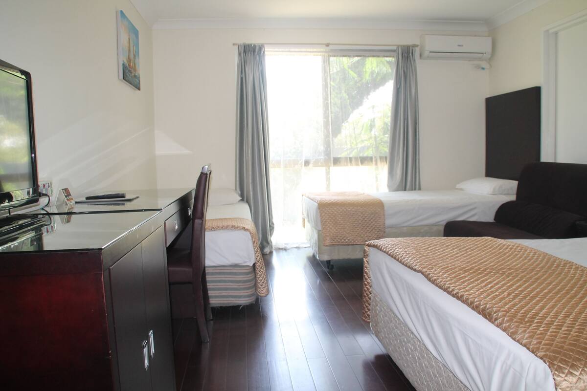A hotel room with a desk, two beds, a chair, and a window with curtains.