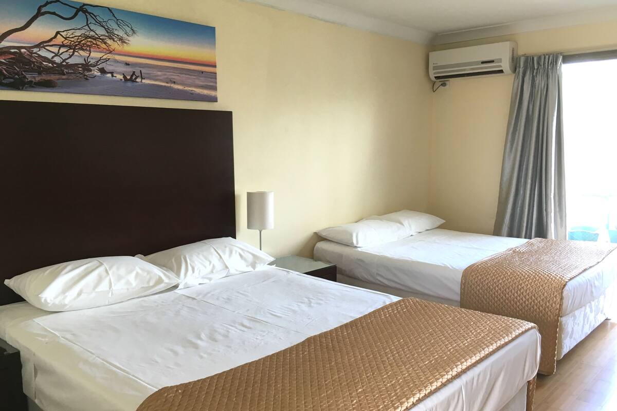 A hotel room with two beds, a painting of a sunset, an air conditioner, and a window with curtains.