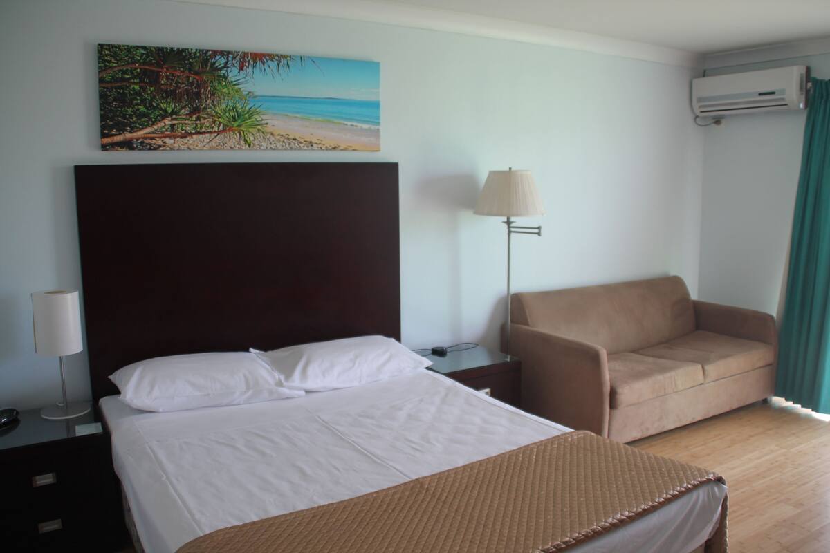 A hotel room with a bed, a sofa, a painting of a beach, a lamp, and an air conditioning unit.